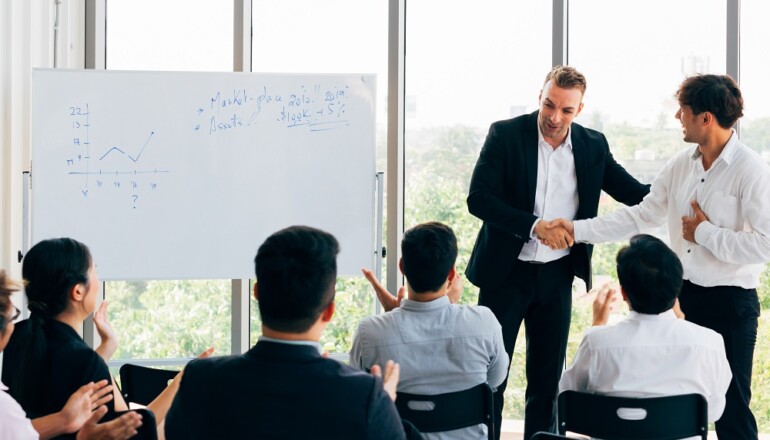 Caucasian business manager introduces new Asian businessman in front of seminar meeting with many employees listening in company office setting